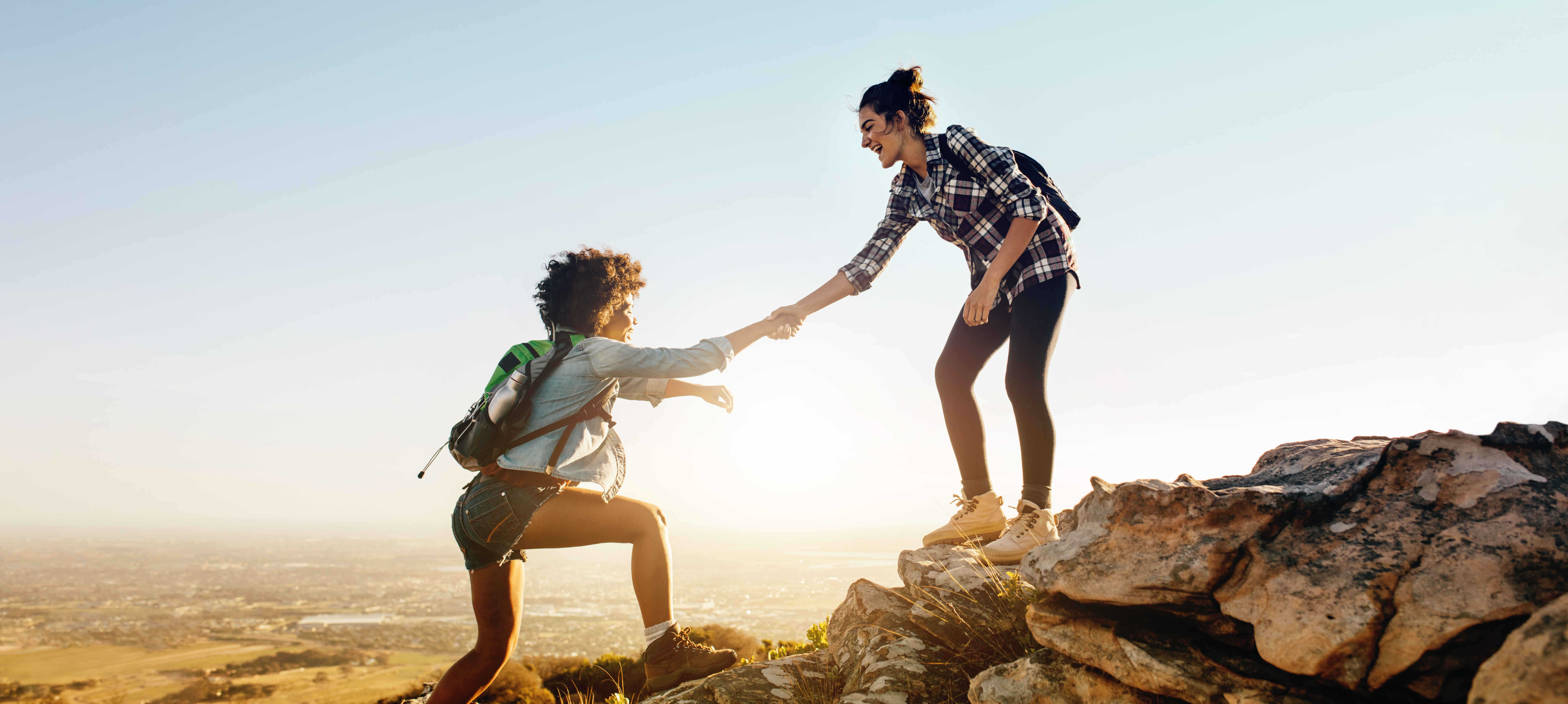 One young woman extending her hand to help another young woman up a rocky cliff.