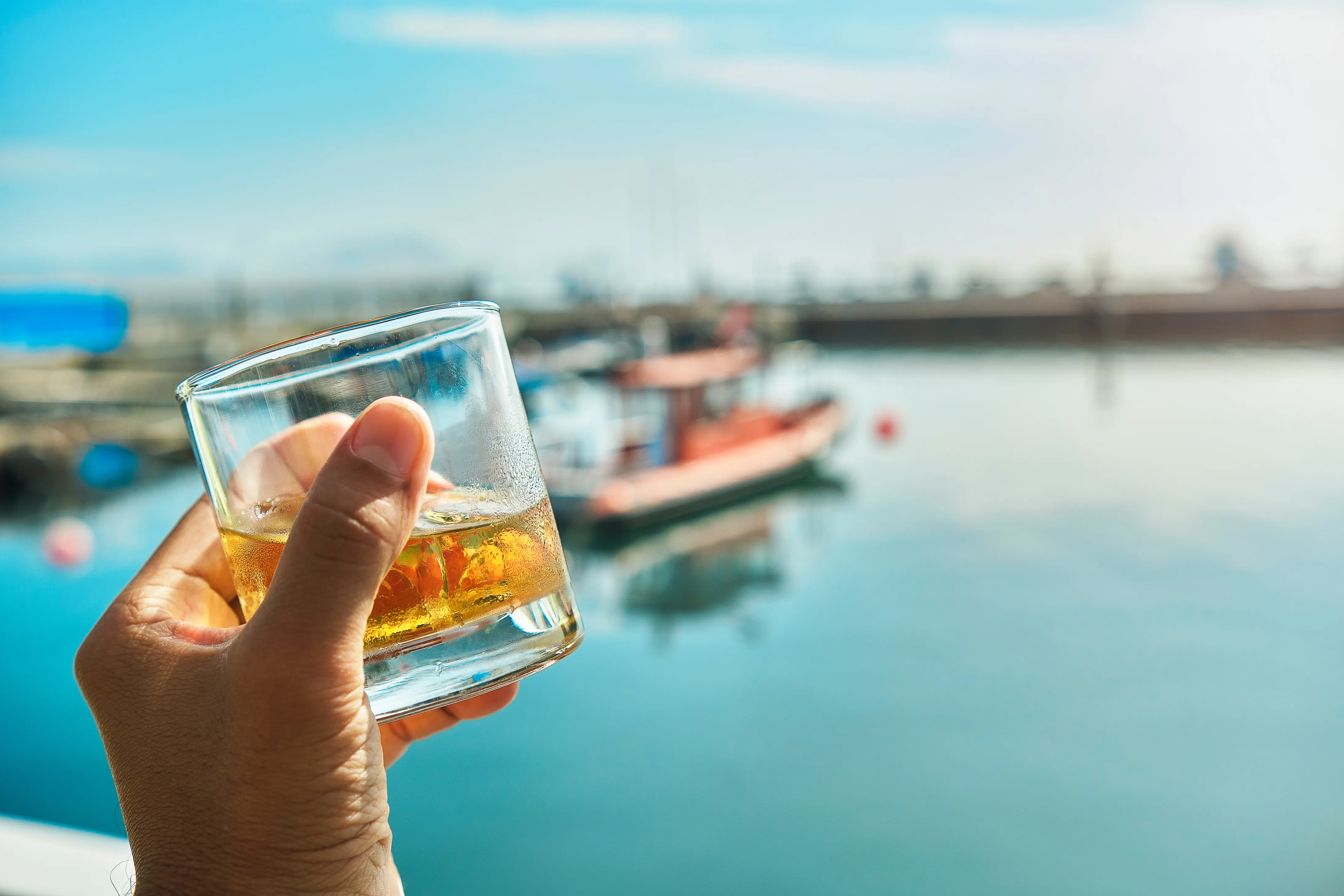 Hand holding a lowball glass with some brown liquor and ice over a summer marina background.