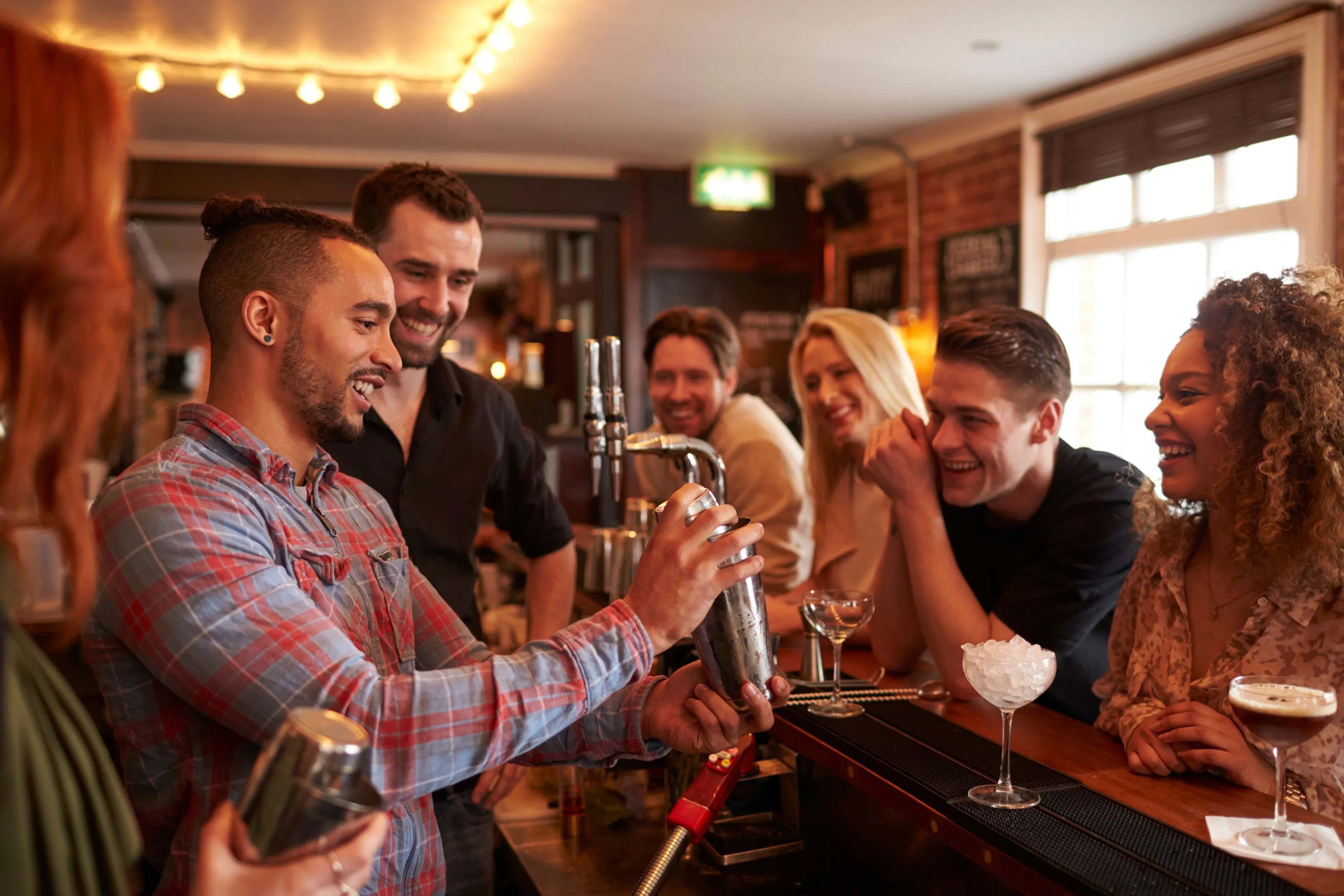 Group of friends watch on as a young man is using a cocktail shaker with guidance from a bartender