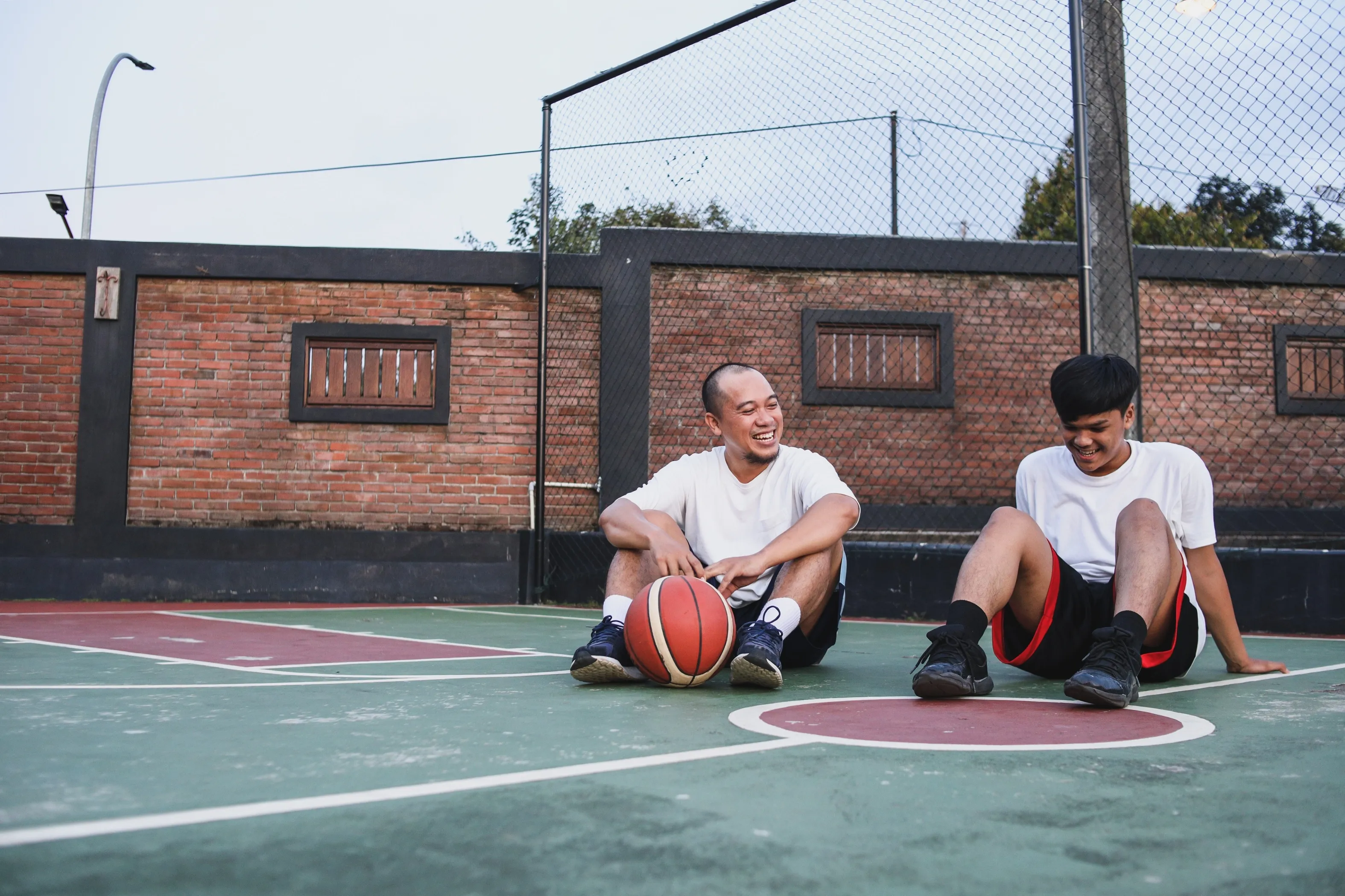 Two male friends, one older and one younger, smiling after a game of basketball.